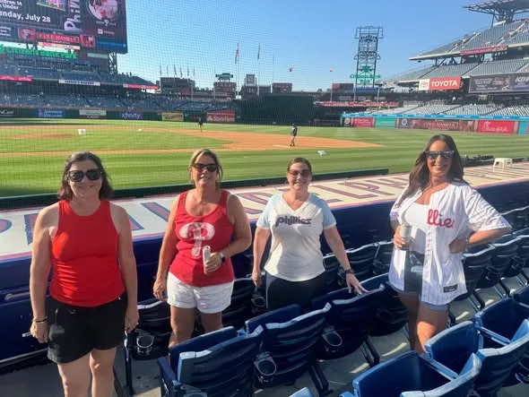 Four women wearing Phillies gear stand together in front row seats overlooking the sunny baseball field at Citizens Bank Park.