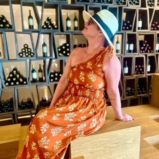 A woman in an orange floral dress and sun hat sits before a geometric wine display wall.