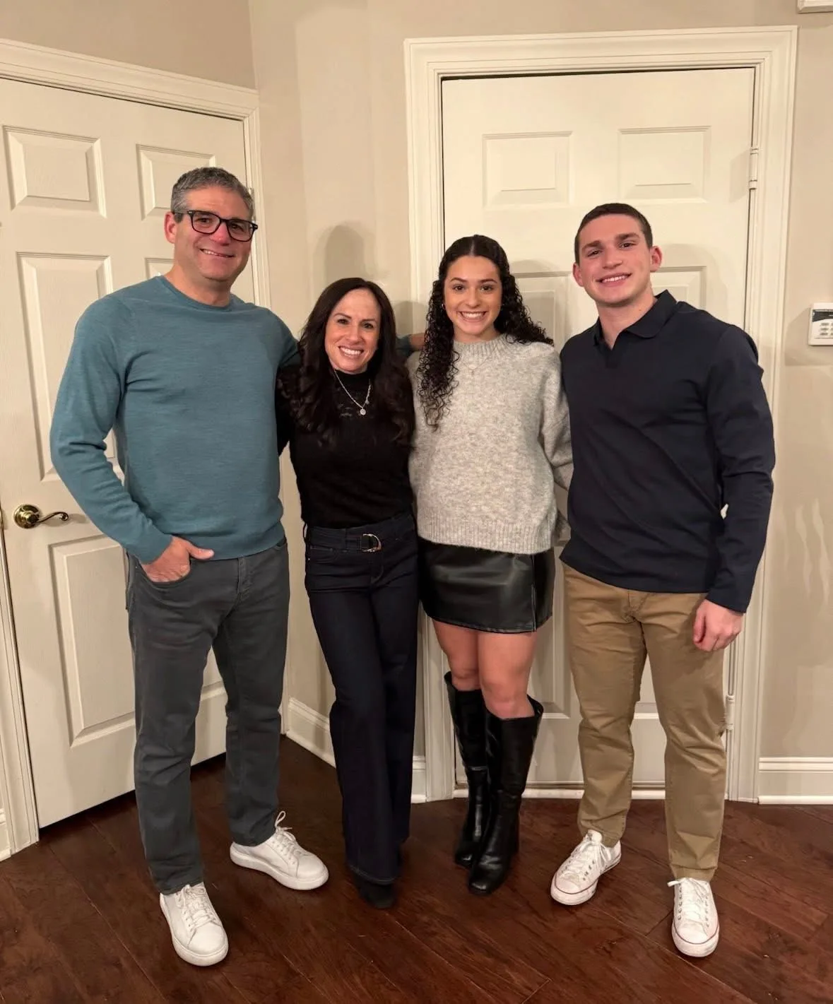 A smiling family of four stands together in casual attire inside a home with white paneled doors.
