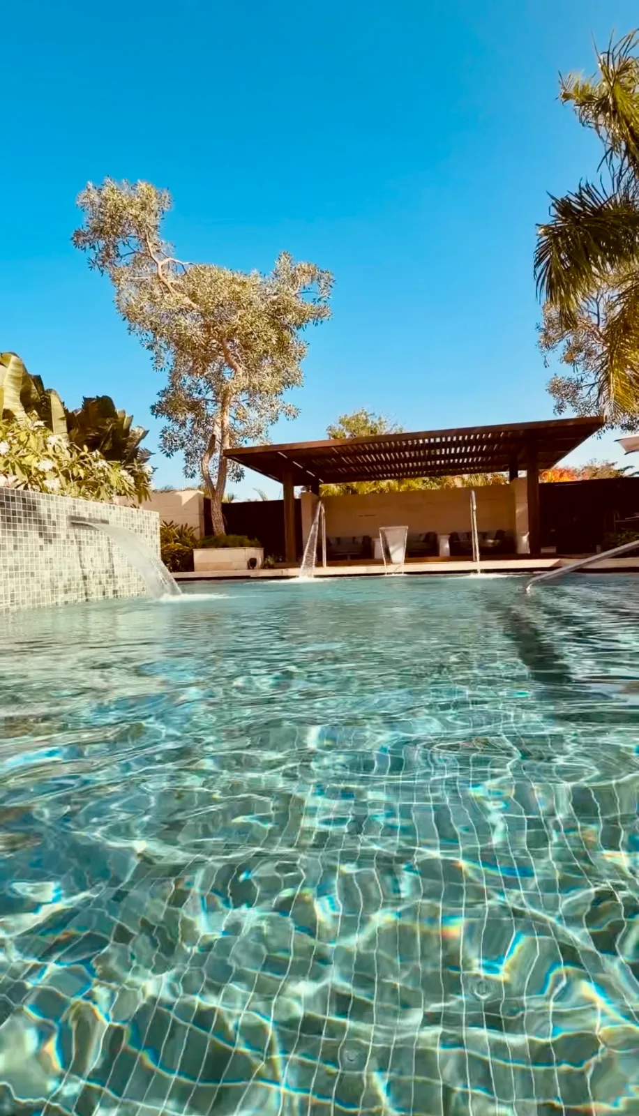 A tiled swimming pool with fountain spouts sits before a shaded wooden cabana framed by tropical trees.