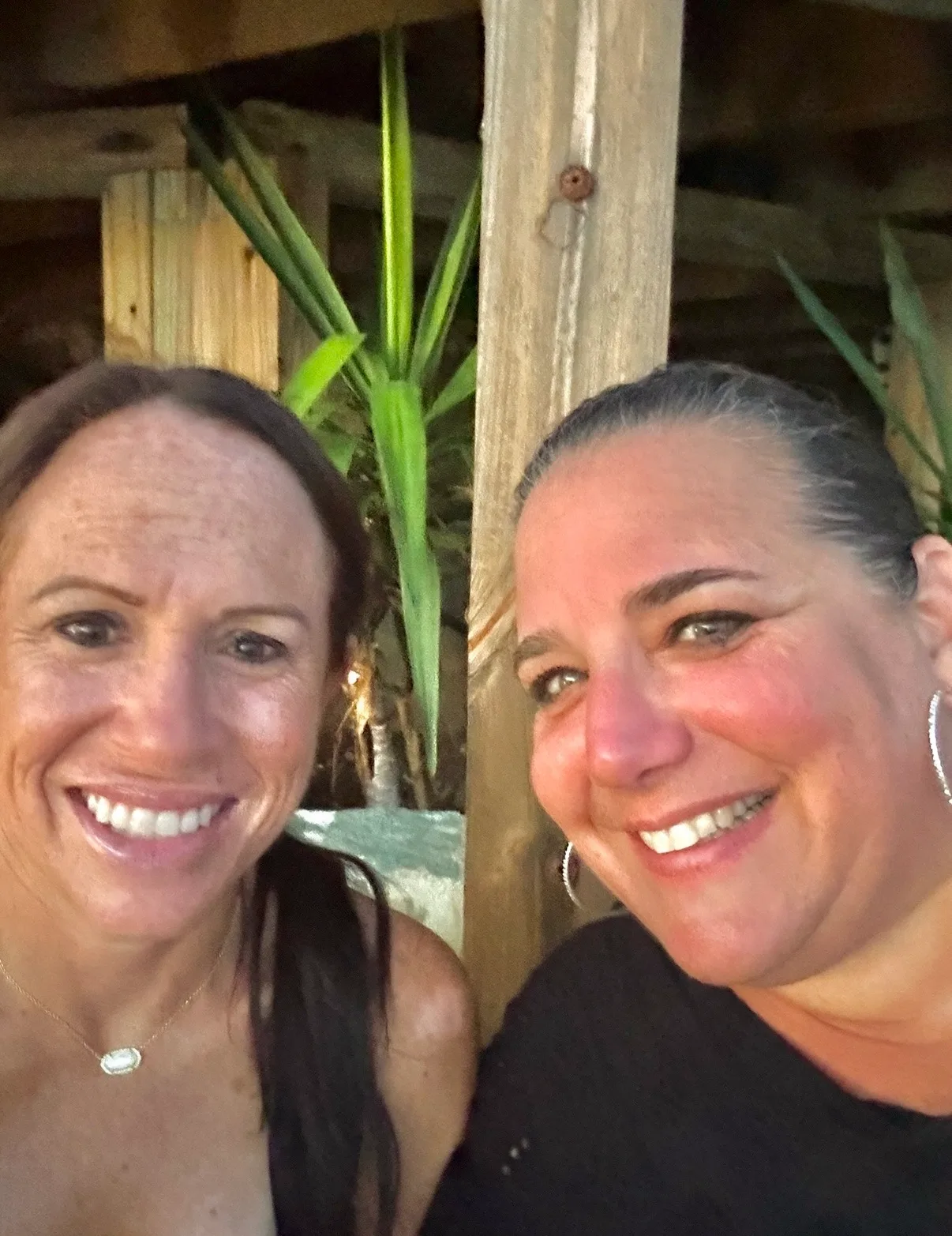 Two smiling women pose together outdoors against a rustic wooden beam and green tropical plants.