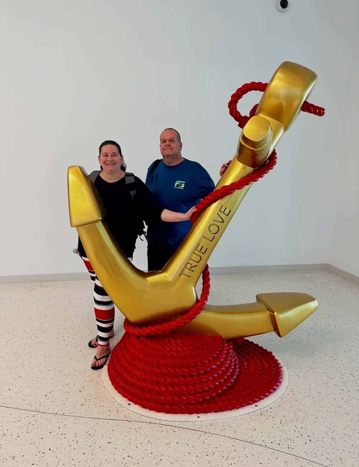 A smiling man and woman pose behind a large golden anchor sculpture wrapped in red rope inside a bright hall.