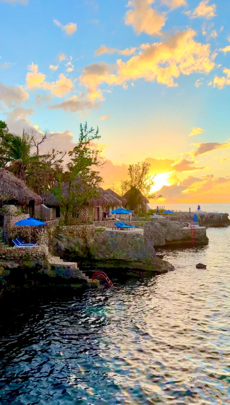 Thatched-roof stone villas with blue umbrellas overlook the ocean from rocky cliffs during a golden sunset in Jamaica.