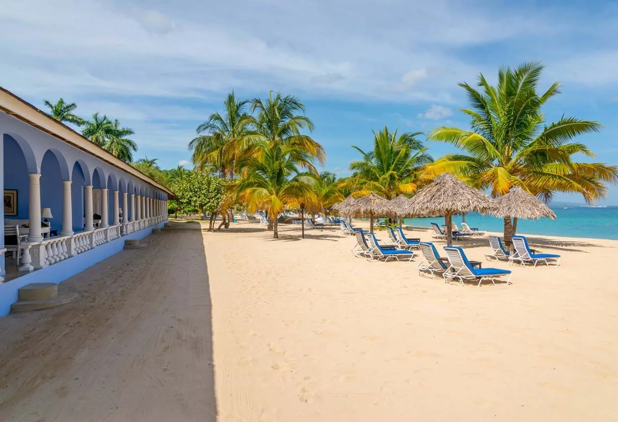 A blue colonnaded verandah opens onto a sandy beach lined with palm trees, thatched umbrellas, and sun loungers in Jamaica.