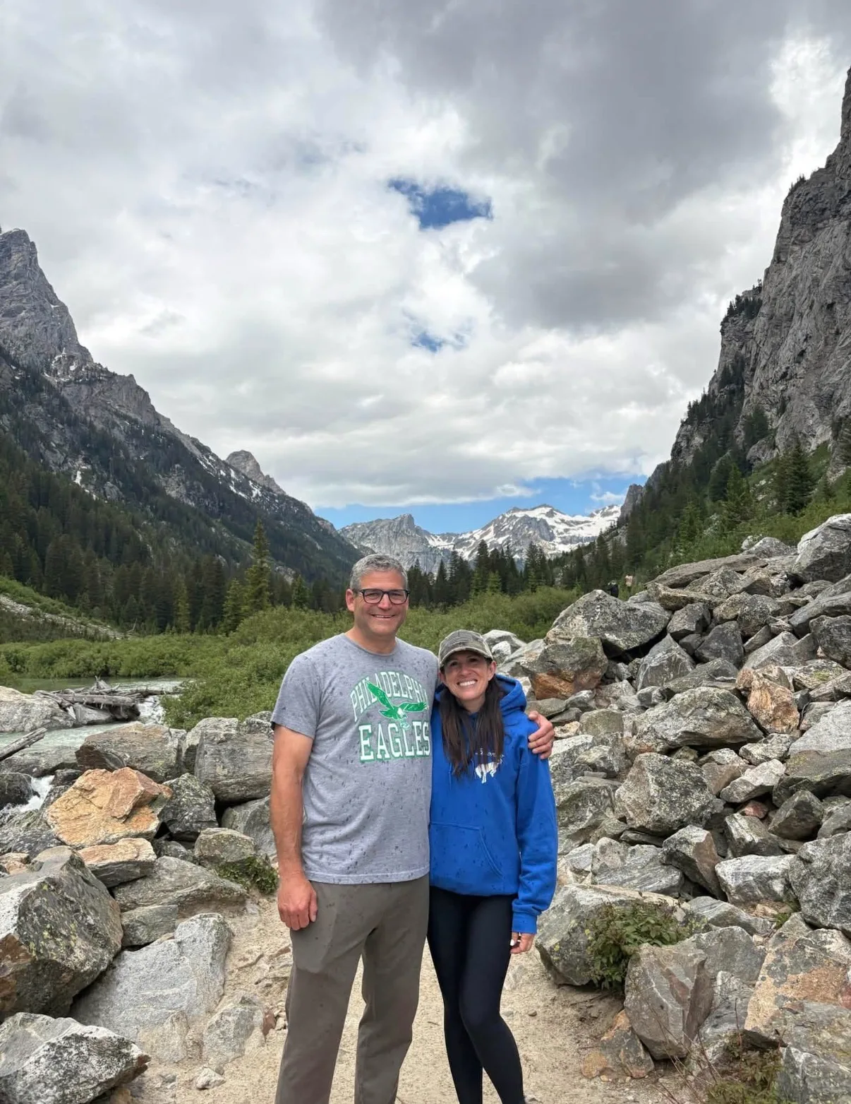 A smiling couple stands on a rocky trail in a rugged mountain valley bordered by evergreen forests and steep peaks.