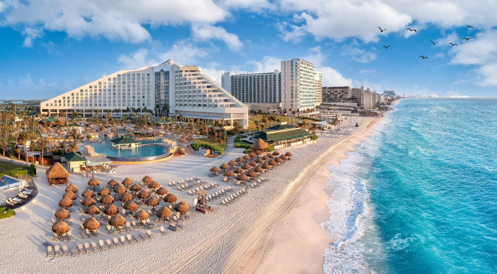 A pyramid-style beachfront resort overlooks swimming pools and thatched palapas along the turquoise ocean in Cancún, Mexico.