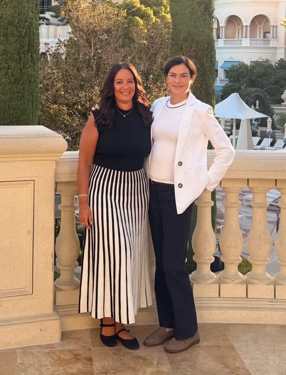 Two smiling women pose together on a stone balustrade terrace overlooking resort gardens and Mediterranean-style architecture.