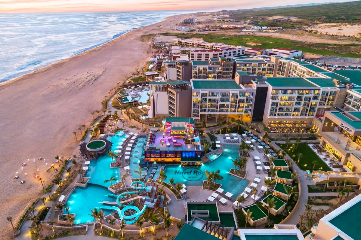 A beachfront resort complex features illuminated swimming pools and waterslides beside a wide sandy coastline in Los Cabos at dusk.