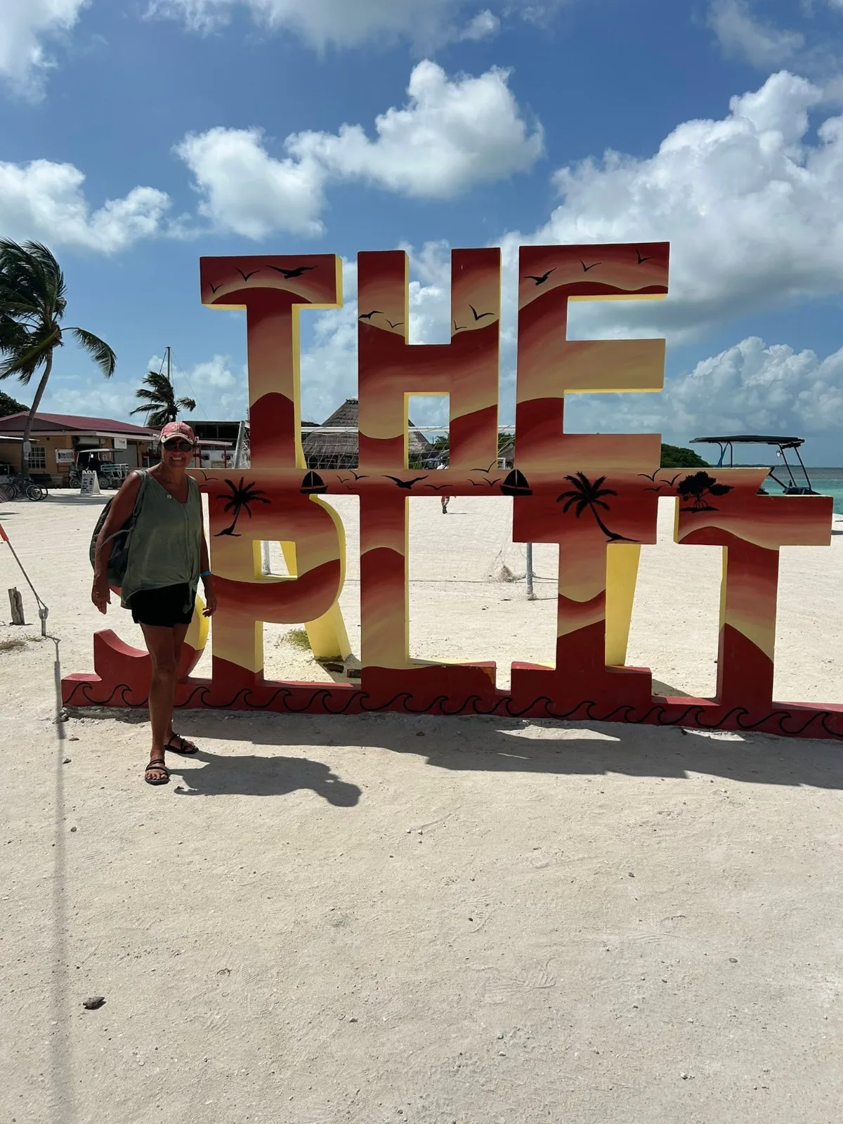 A woman stands beside a large colorful sign reading The Split on a sandy tropical beach under a partly cloudy sky.