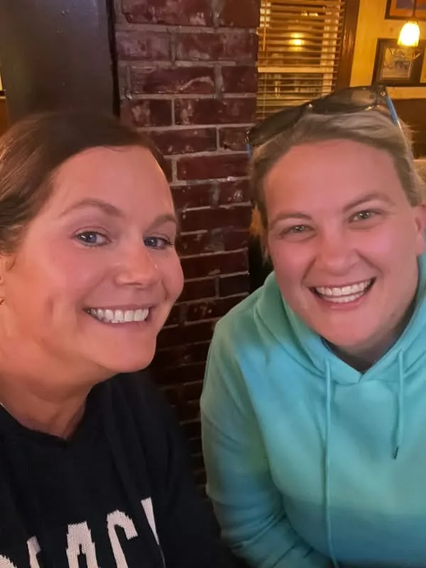 Two smiling women pose together inside a restaurant beside an exposed brick wall with warm background lighting.