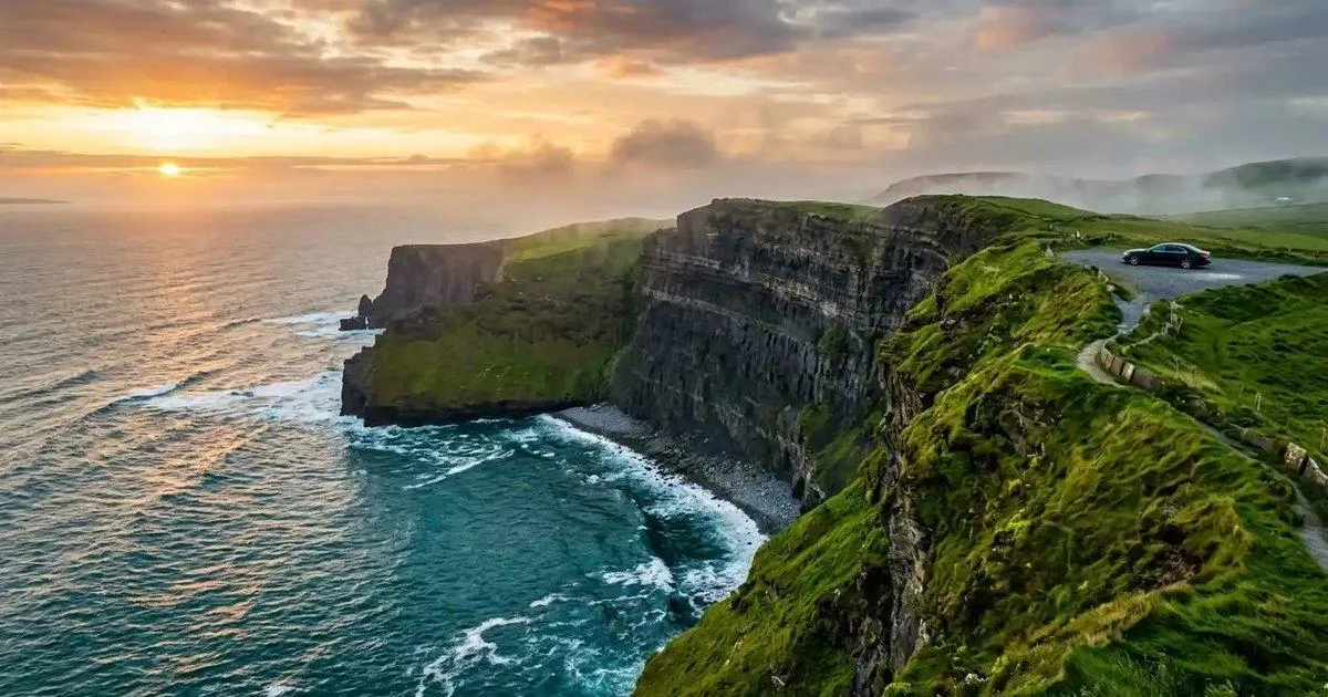 Green coastal cliffs rise above crashing ocean waves at sunset along the Cliffs of Moher in Ireland.