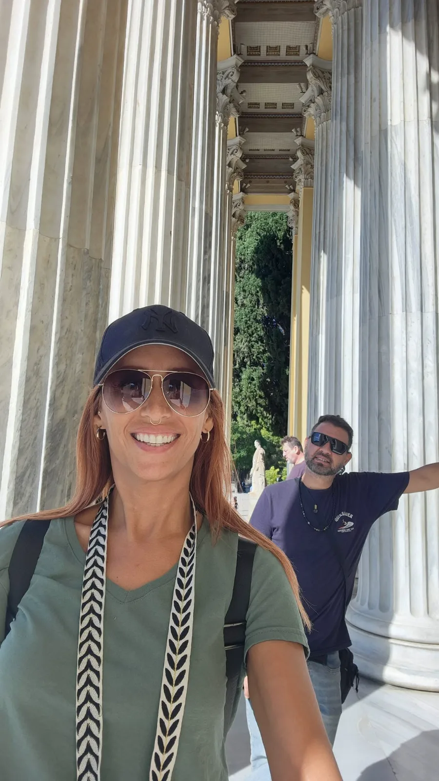 A smiling woman wearing sunglasses poses for a selfie with a companion between towering classical marble columns in Athens.