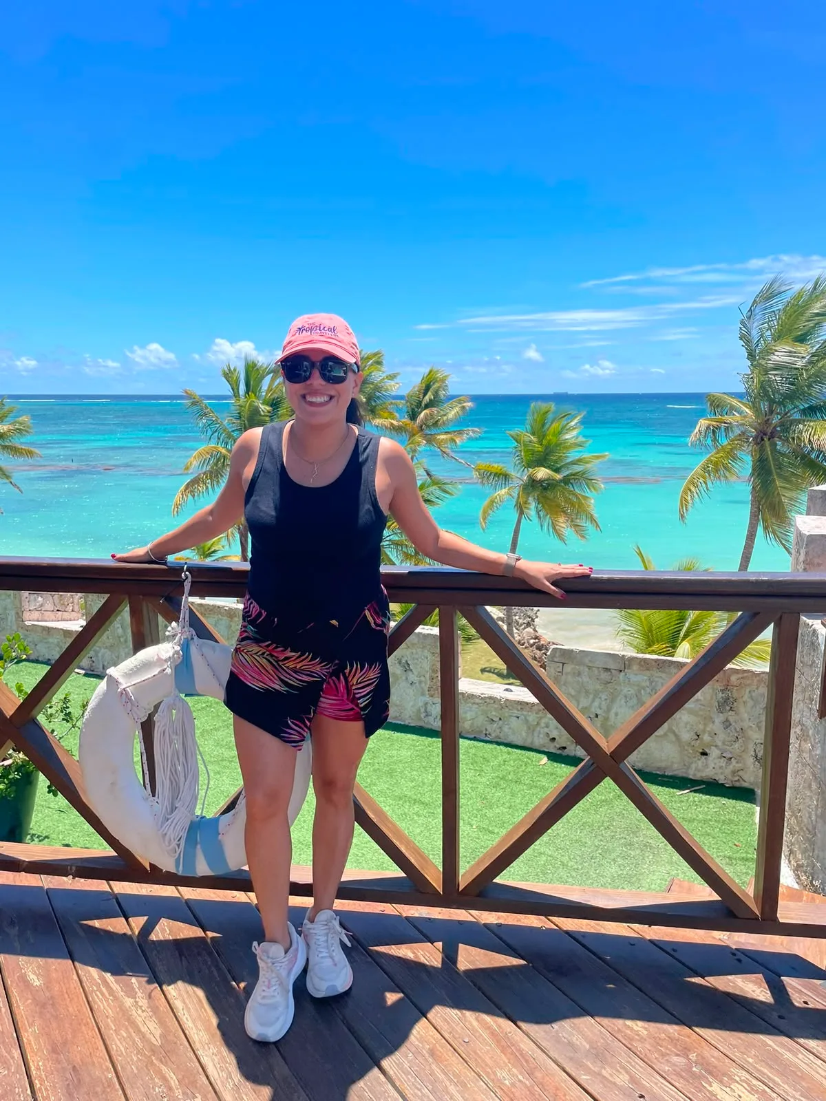 A smiling woman wearing sunglasses stands on a wooden deck overlooking palm trees and turquoise tropical ocean waters.