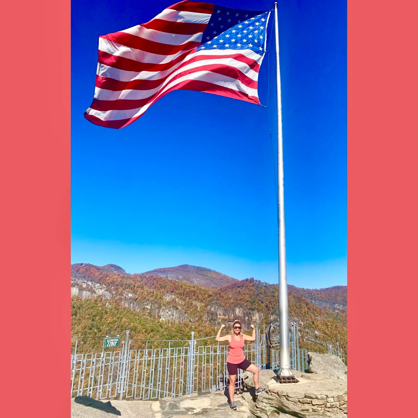 A large American flag waves above a woman posing on a rocky mountain overlook against forested autumn hills.