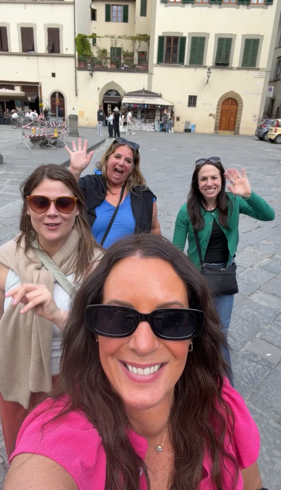 Four smiling women wave while taking a selfie together in a paved European city square lined with historic buildings.