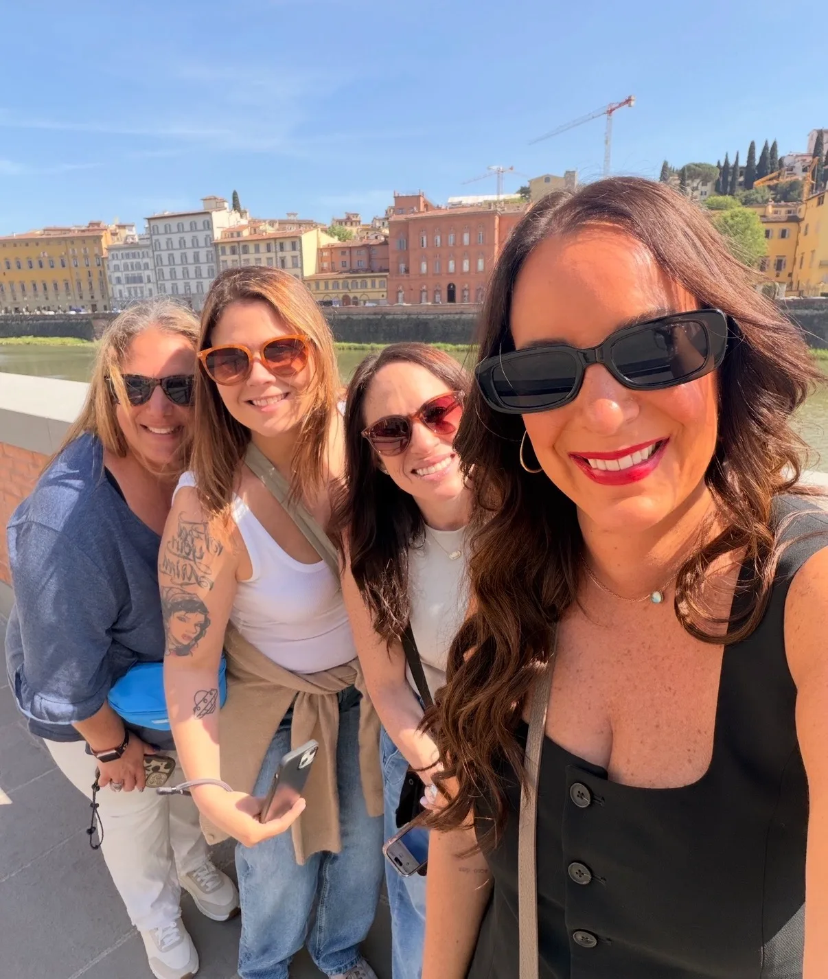 Four women wearing sunglasses smile for a selfie along a sunny riverside promenade lined with historic Italian buildings.