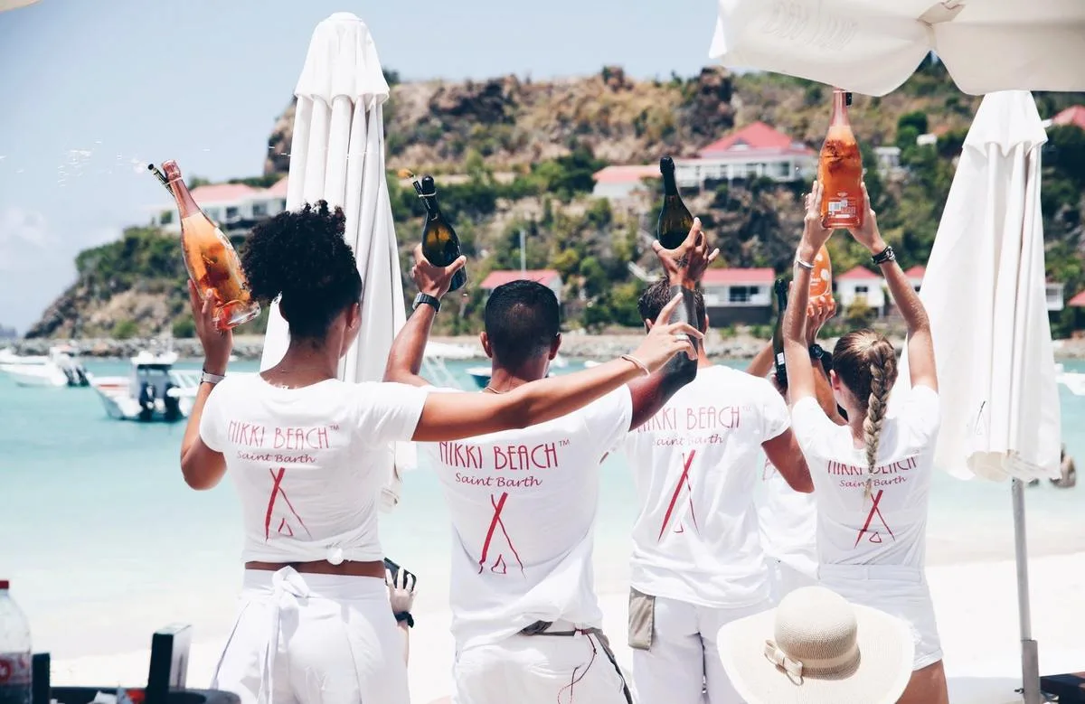Waitstaff in matching white uniforms raise champagne bottles overhead along a sunny beach club in Saint Barthélemy.