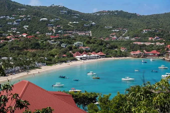 Motorboats anchor in a turquoise bay bordered by a sandy beach and green hillsides dotted with villas in St. Barts.