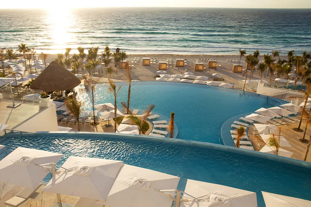 Tiered infinity pools lined with white umbrellas overlook a sandy beach and the ocean at a resort in Cancun.