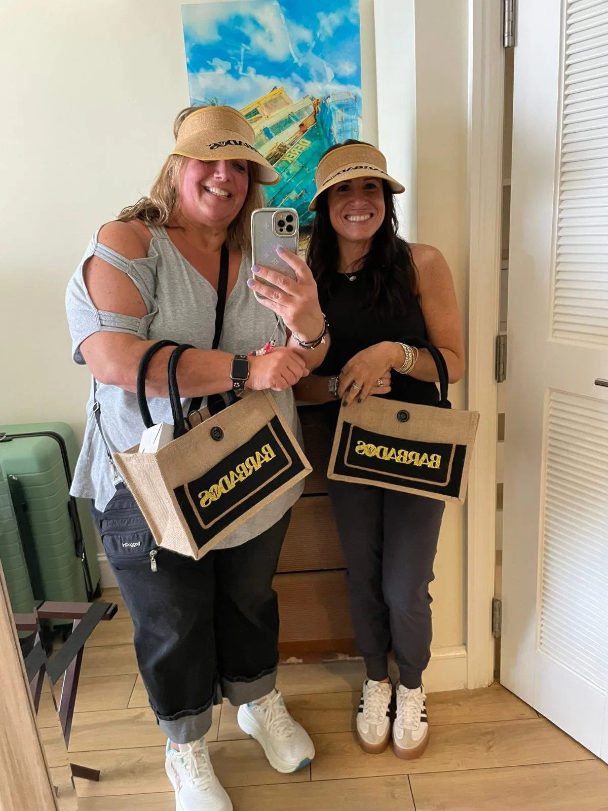 Two smiling women take a mirror selfie wearing matching straw visors and holding Barbados tote bags inside a resort room.