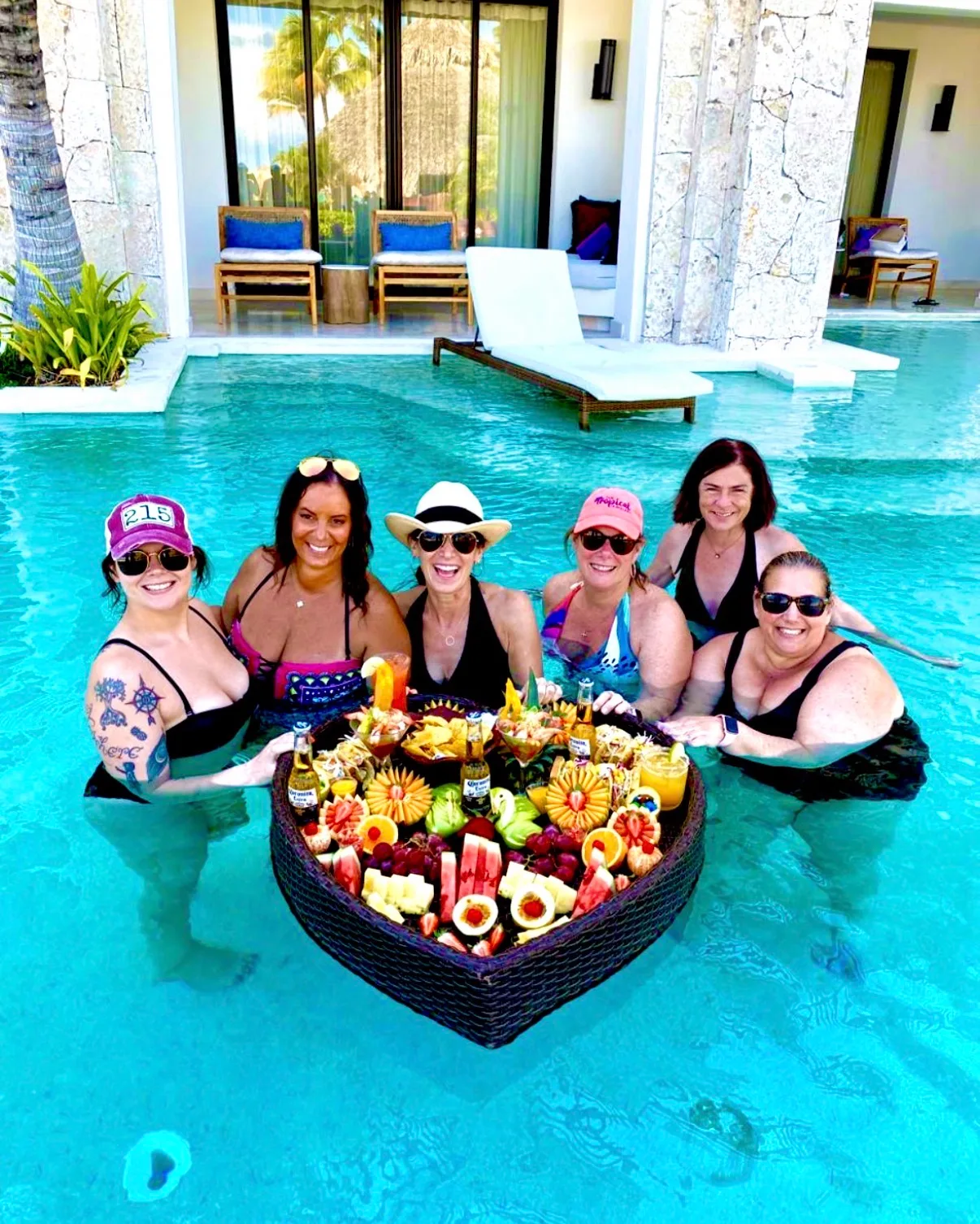 Six women in swimwear gather around a floating tray of fruit and drinks in a turquoise resort swimming pool.