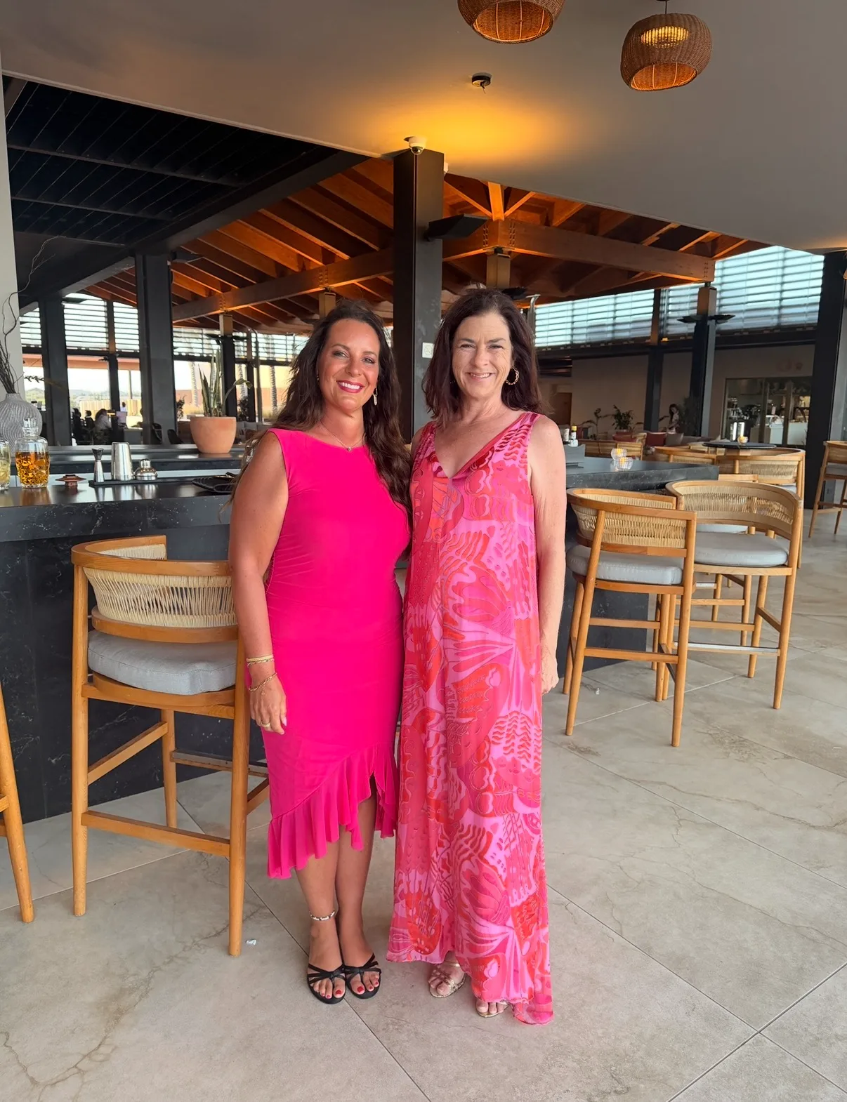 Two women in bright pink dresses smile together in an open-air resort lounge beside a modern bar.