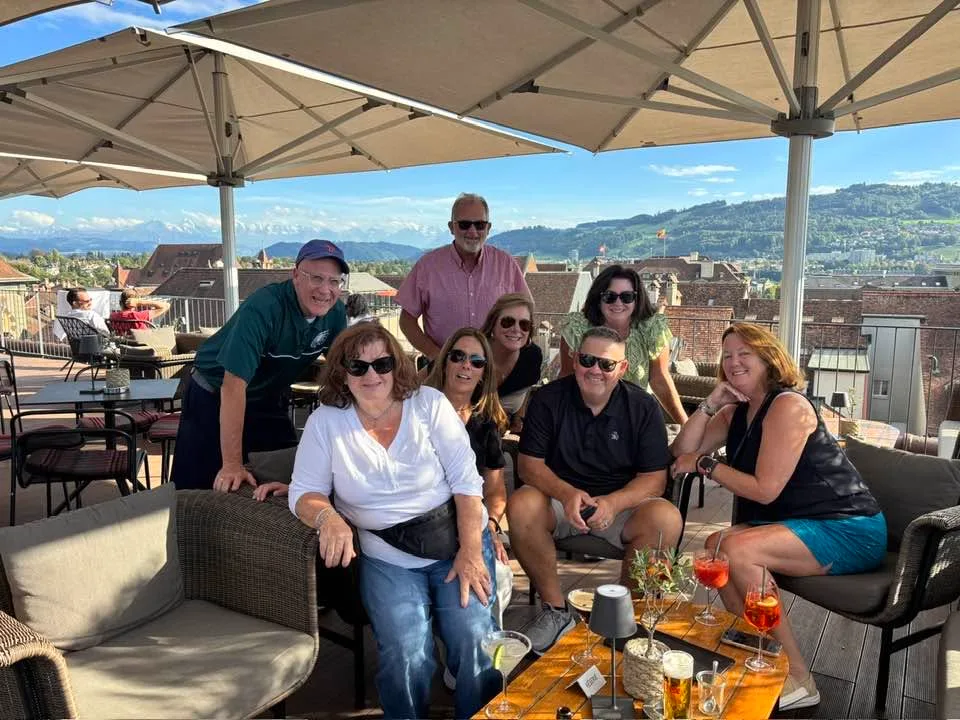 A group of friends smiles together on a sunny rooftop terrace with drinks, overlooking historic European rooftops and distant mountains.