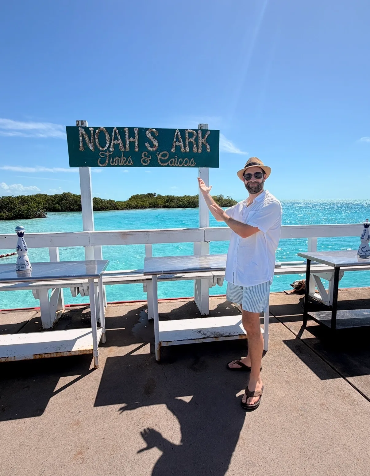 A man in a sun hat poses beside a Noah's Ark sign overlooking turquoise water in Turks and Caicos.