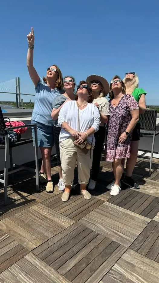 A group of women wearing solar eclipse glasses look up toward the clear sky from a wooden rooftop terrace.