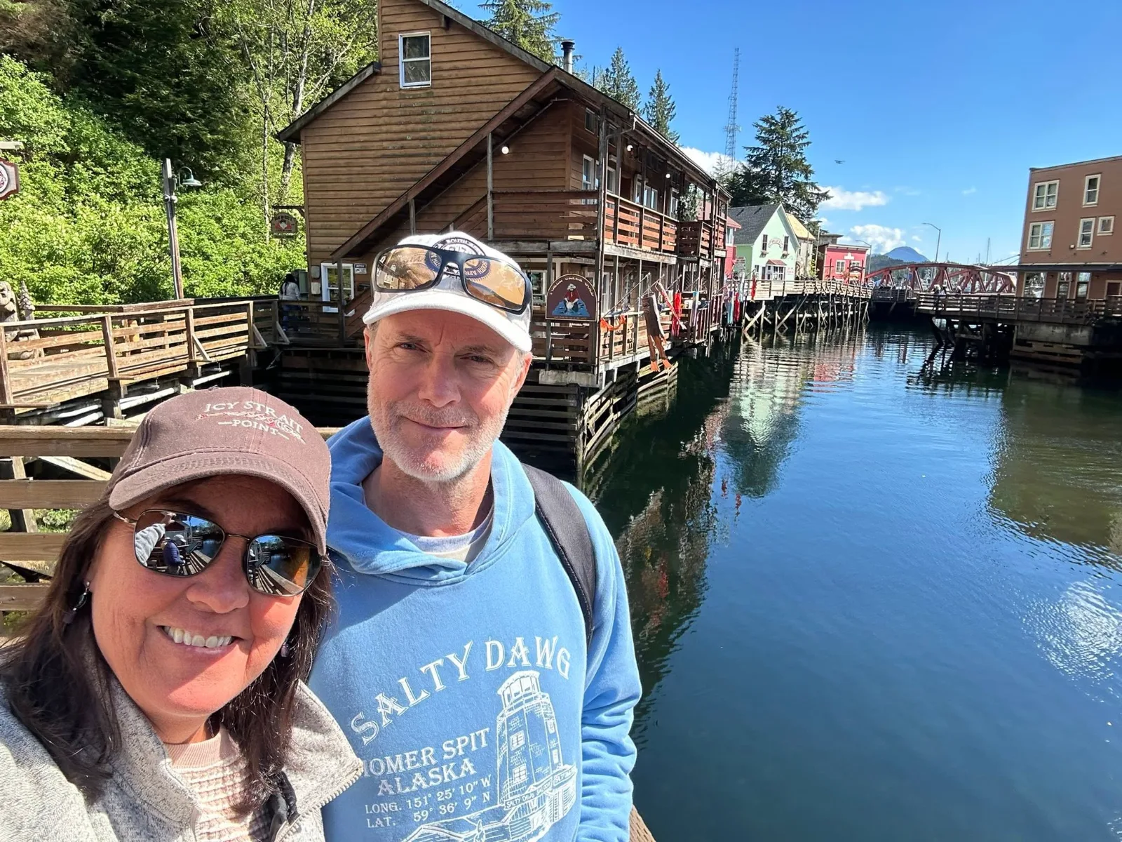 A smiling couple takes a selfie beside historic overwater wooden buildings along Creek Street in Ketchikan, Alaska.