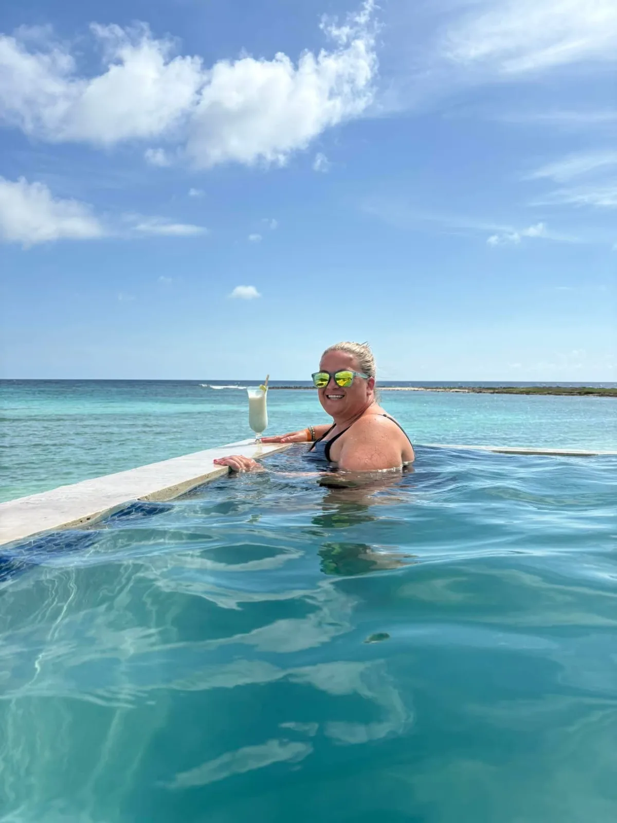 A smiling woman in sunglasses relaxes at the edge of an infinity pool with a cocktail overlooking the turquoise sea.