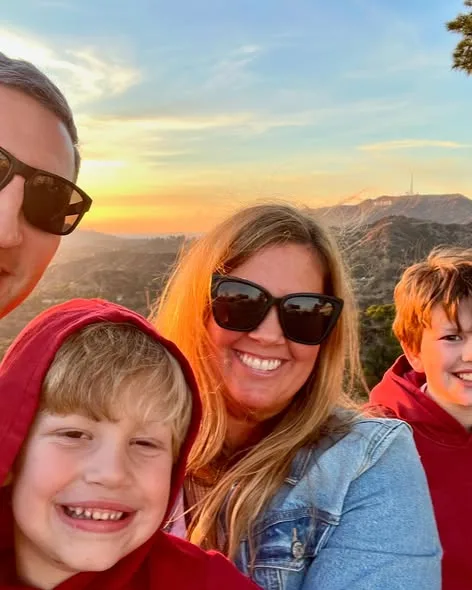 A smiling family poses for an outdoor selfie at sunset with the Hollywood Sign visible in the background.