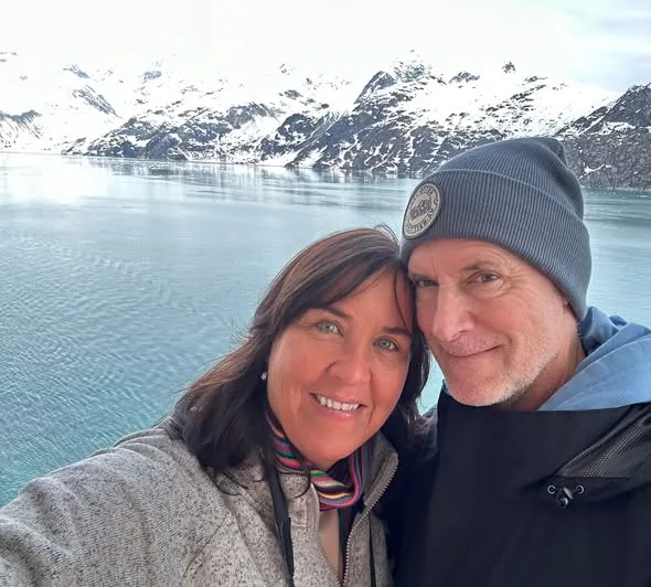 A smiling couple dressed in warm outerwear poses together before snow-covered mountains and calm fjord waters.