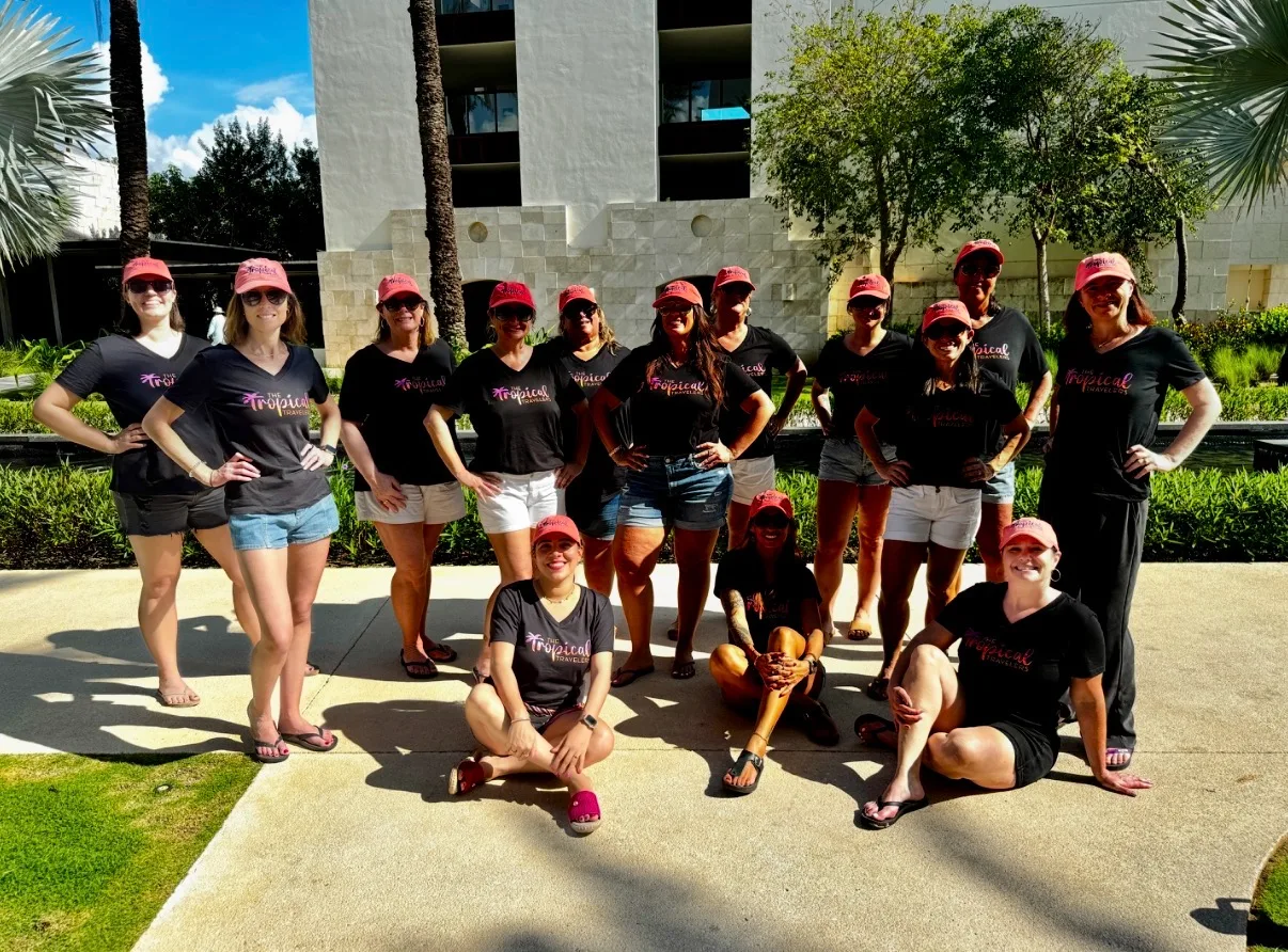 A group of women in matching black shirts and pink caps poses outdoors at a sunny tropical resort courtyard.