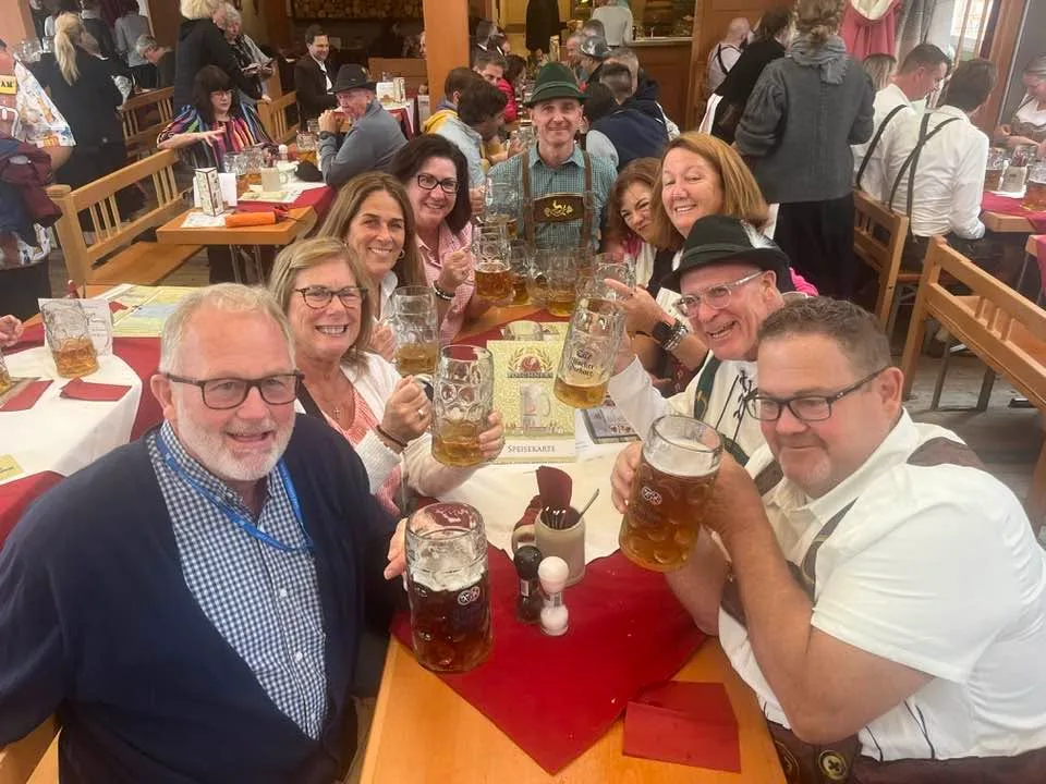 A cheerful group of travelers raises large beer steins at a crowded table in a traditional Bavarian beer hall.