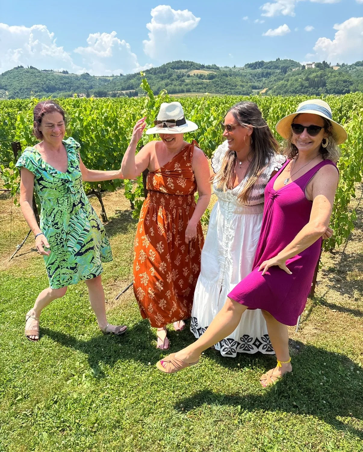 Four women in colorful summer dresses and sun hats pose along a sunny vineyard framed by rolling green hills.