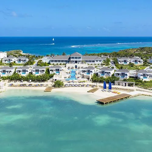 White beachfront resort villas and swimming pools overlook a calm turquoise bay and white sand beach in Antigua.