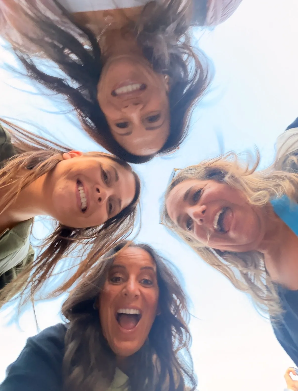 Four smiling women look down together at the camera against a bright daylight sky.
