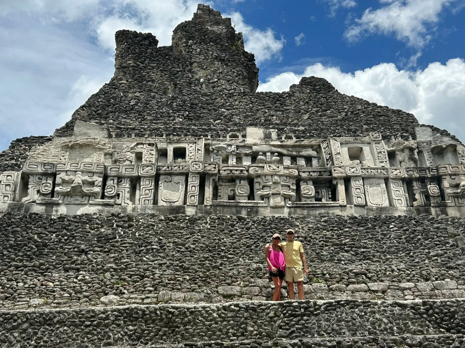 A couple stands on the stone steps of an ancient Maya pyramid beneath intricate carved friezes under a bright sky.