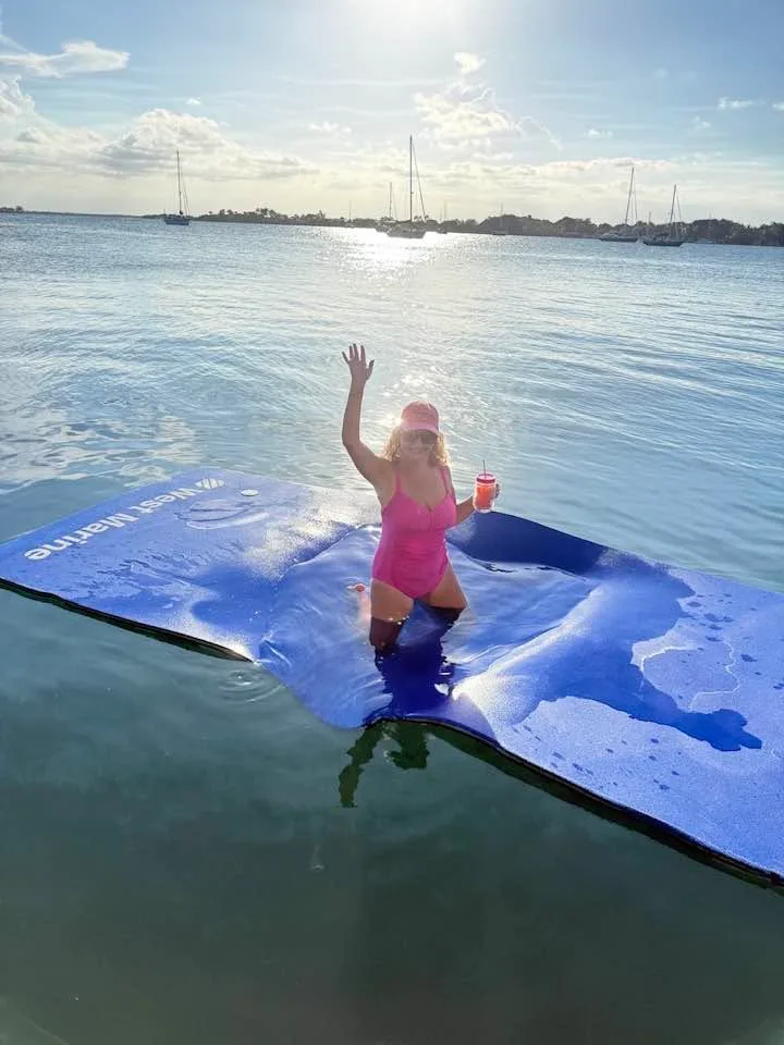 A woman in a pink swimsuit waves from a blue floating mat on calm coastal waters with sailboats anchored nearby.