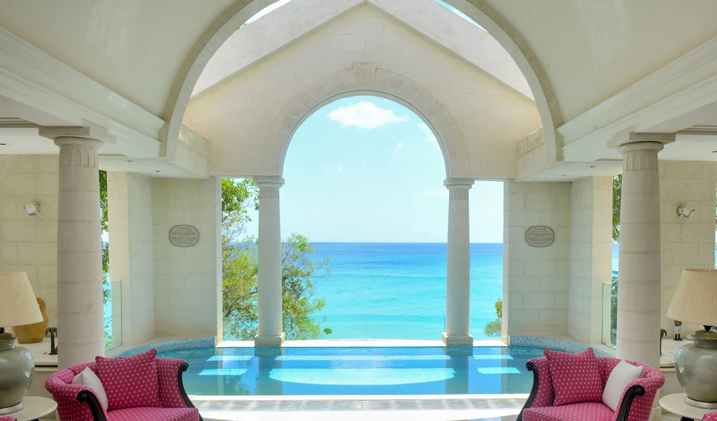 A stone-arched spa pool flanked by pink armchairs overlooks the turquoise Caribbean Sea in Barbados.