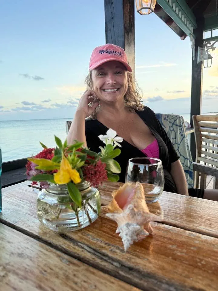 A smiling woman in a pink cap sits at a seaside deck table decorated with flowers and a conch shell at sunset.