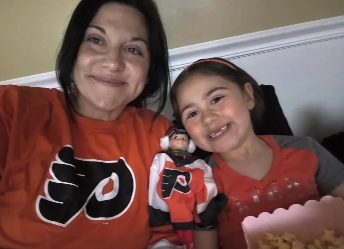 A smiling woman and child wearing Philadelphia Flyers shirts pose indoors with a small dressed doll and popcorn.