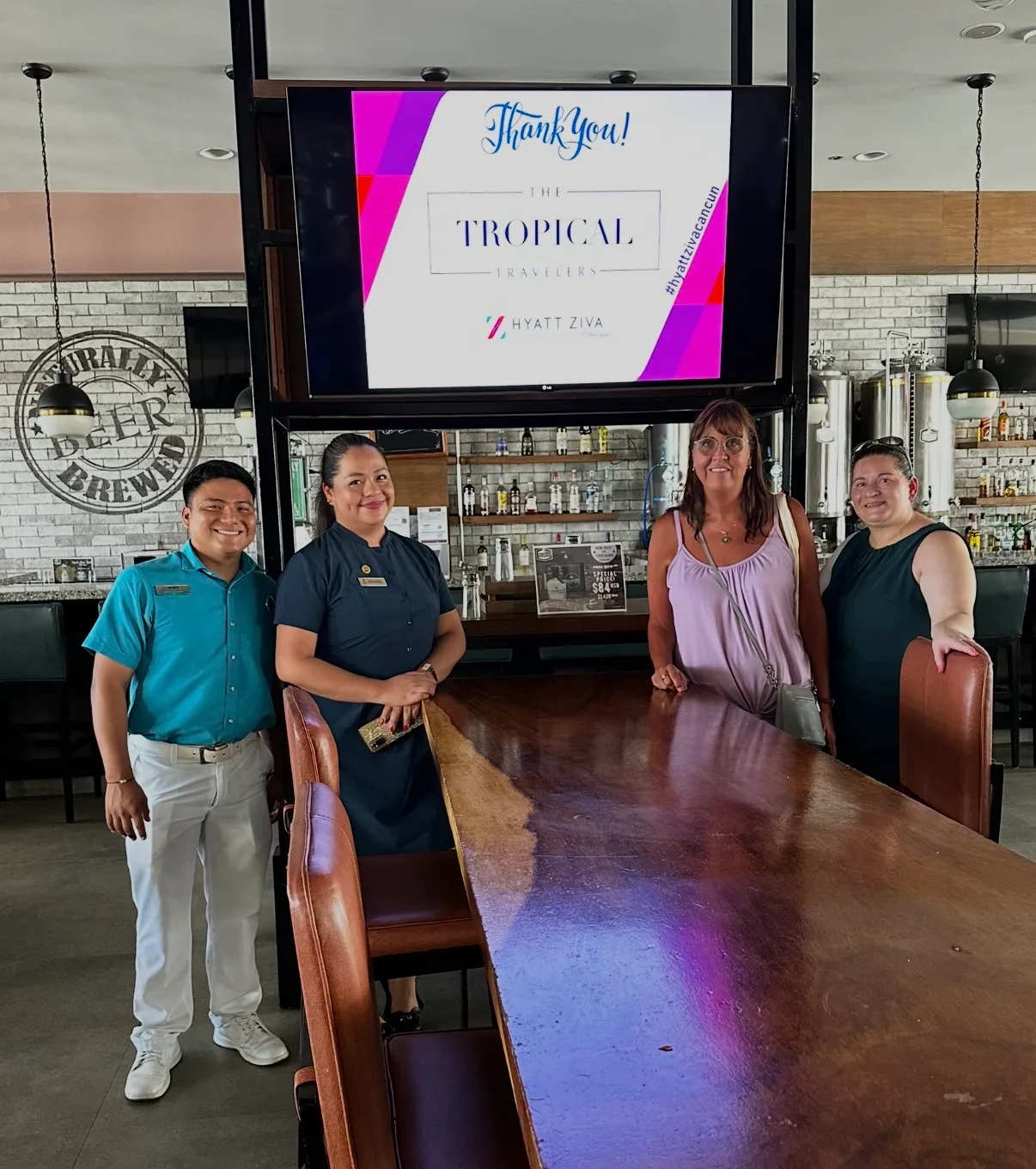 Resort staff and guests stand smiling beside a polished wooden table in the microbrewery bar at Hyatt Ziva Cancun.