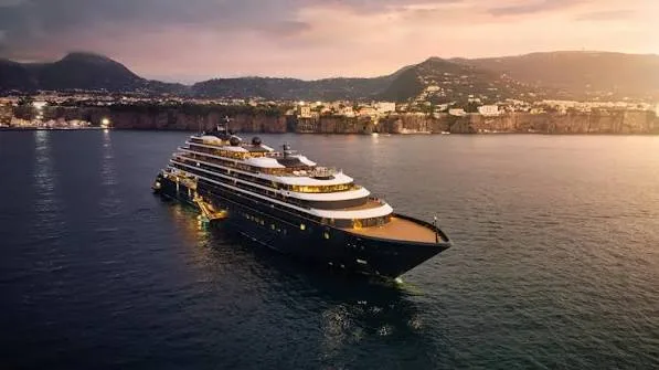 An illuminated luxury cruise yacht navigates calm waters off a steep, mountainous coastline during golden hour at dusk.