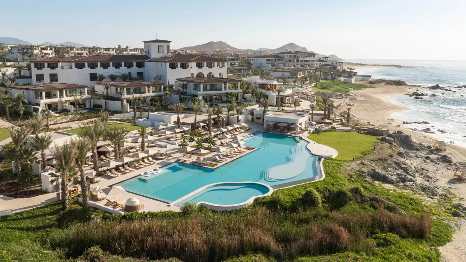 White resort buildings and a tiered oceanfront swimming pool overlook a sandy beach in Los Cabos, Mexico.
