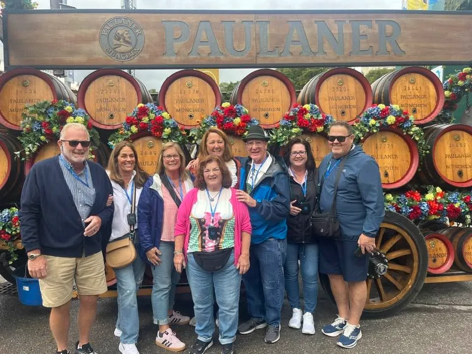 A smiling group of travelers stands together in front of a flower-adorned Paulaner brewery wagon.