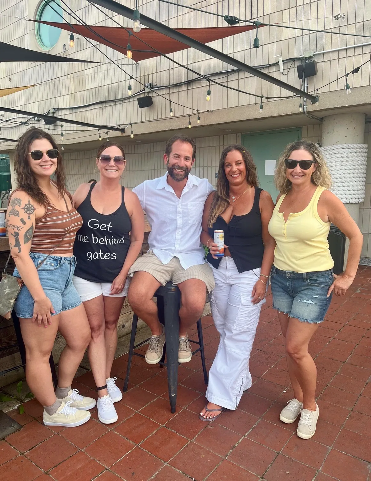Five friends pose together on an outdoor brick patio beneath hanging string lights and shade sails.