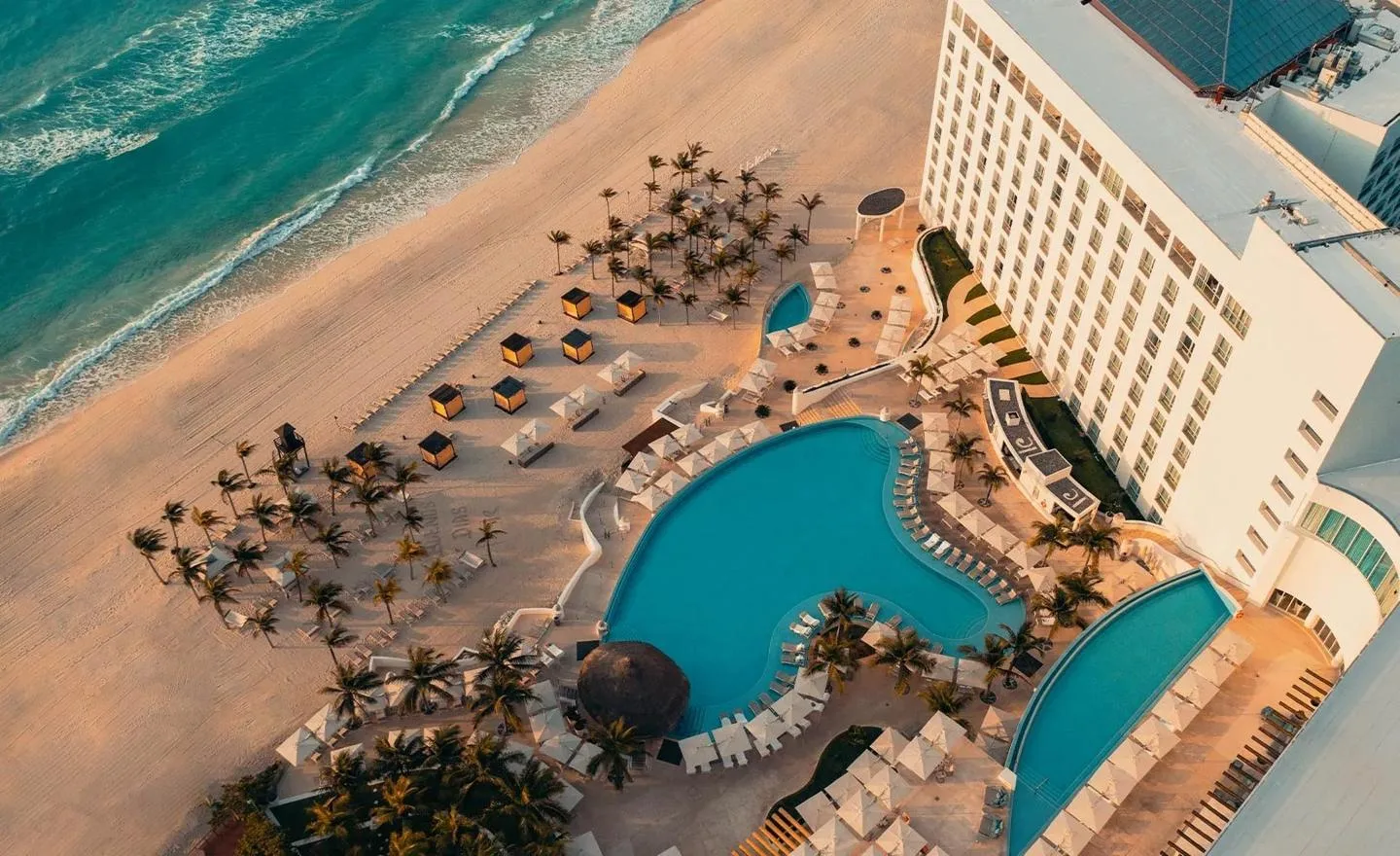 A beachfront resort features curving swimming pools, sun loungers, and palm trees beside the turquoise ocean in Cancún, Mexico.