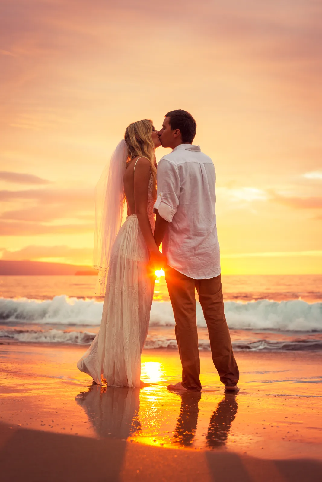 A bride and groom kiss at the water's edge on a sandy beach during a bright ocean sunset.